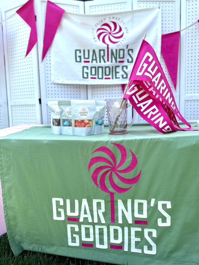May include: A green tablecloth with the text "Guarino's Goodies" and a pink and white swirl lollipop design. Three bags of candy are on the table, each with the text "Guarino's Goodies" and a different color of candy inside.  Pink and white flags with the text "Guarino's" are in the background.