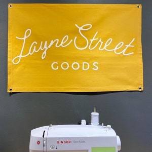 May include: A yellow banner with the text "Layne Street Goods" in white cursive lettering hangs above a white sewing machine with a green digital display. The sewing machine is on a wooden surface.
