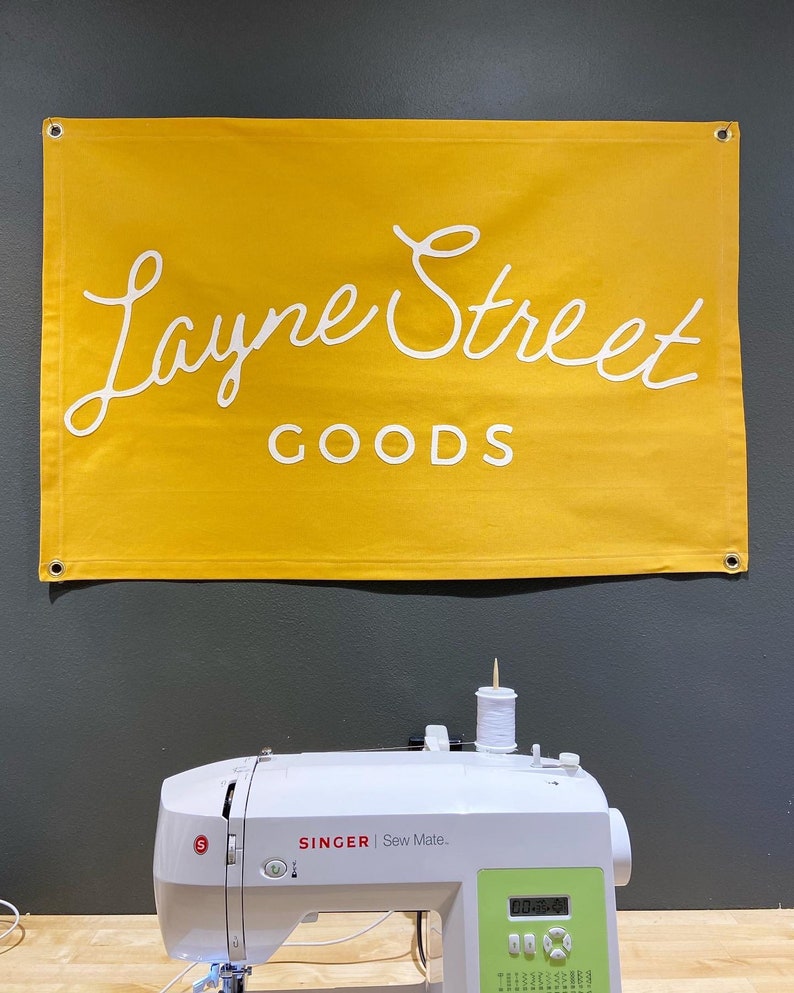 May include: A yellow banner with the text "Layne Street Goods" in white cursive lettering hangs above a white sewing machine with a green digital display. The sewing machine is on a wooden surface.