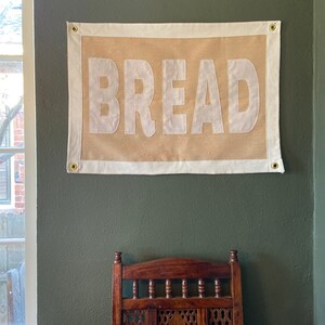 May include: A burlap banner with white text that reads "BREAD" hanging on a green wall. The banner has a white border and brass grommets in the top corners.