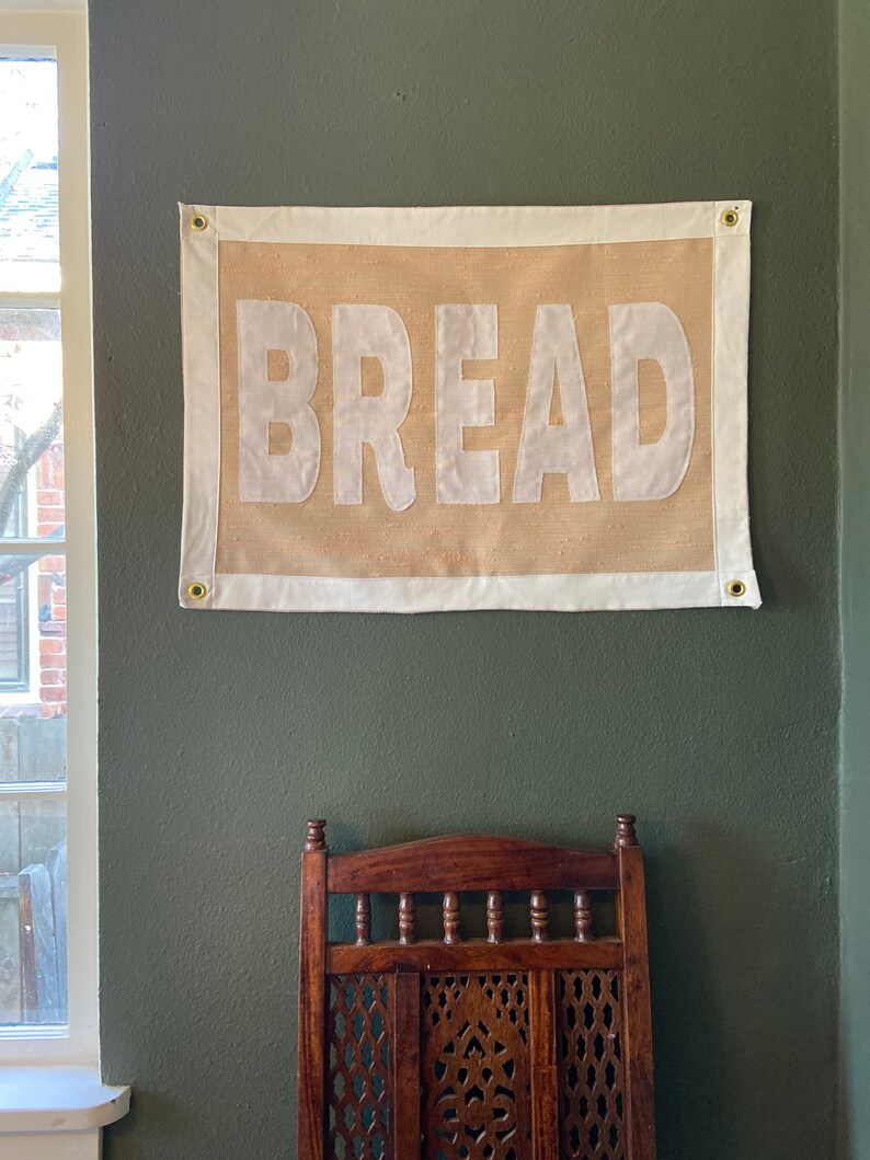 May include: A burlap banner with white text that reads "BREAD" hanging on a green wall. The banner has a white border and brass grommets in the top corners.