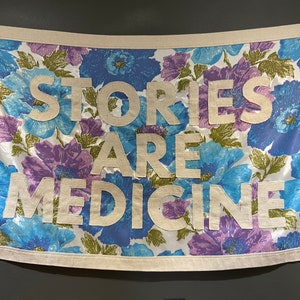May include: A white fabric banner with a floral pattern in shades of blue, purple, and green. The banner has the text "Stories Are Medicine" in white letters.