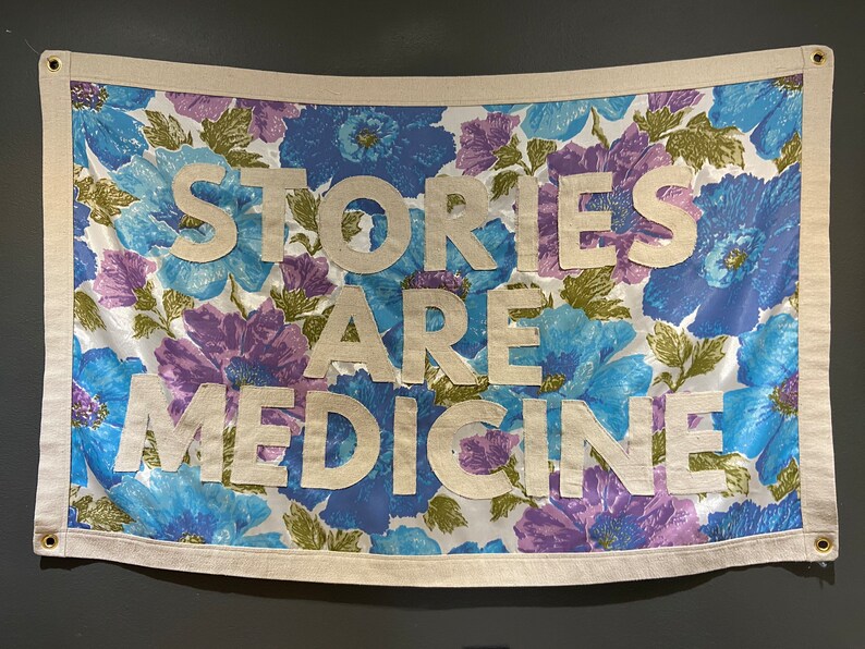 May include: A white fabric banner with a floral pattern in shades of blue, purple, and green. The banner has the text "Stories Are Medicine" in white letters.