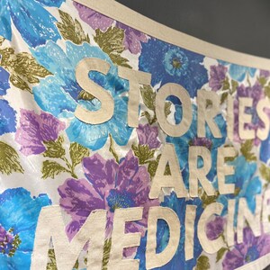 May include: A white fabric banner with a floral pattern in shades of blue and purple. The banner has the text "Stories Are Medicine" written in white fabric letters.