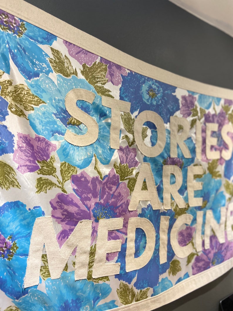 May include: A white fabric banner with a floral pattern in shades of blue and purple. The banner has the text "Stories Are Medicine" written in white fabric letters.