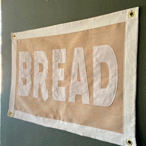 May include: A beige canvas banner with white trim and the word "BREAD" printed in large white letters.