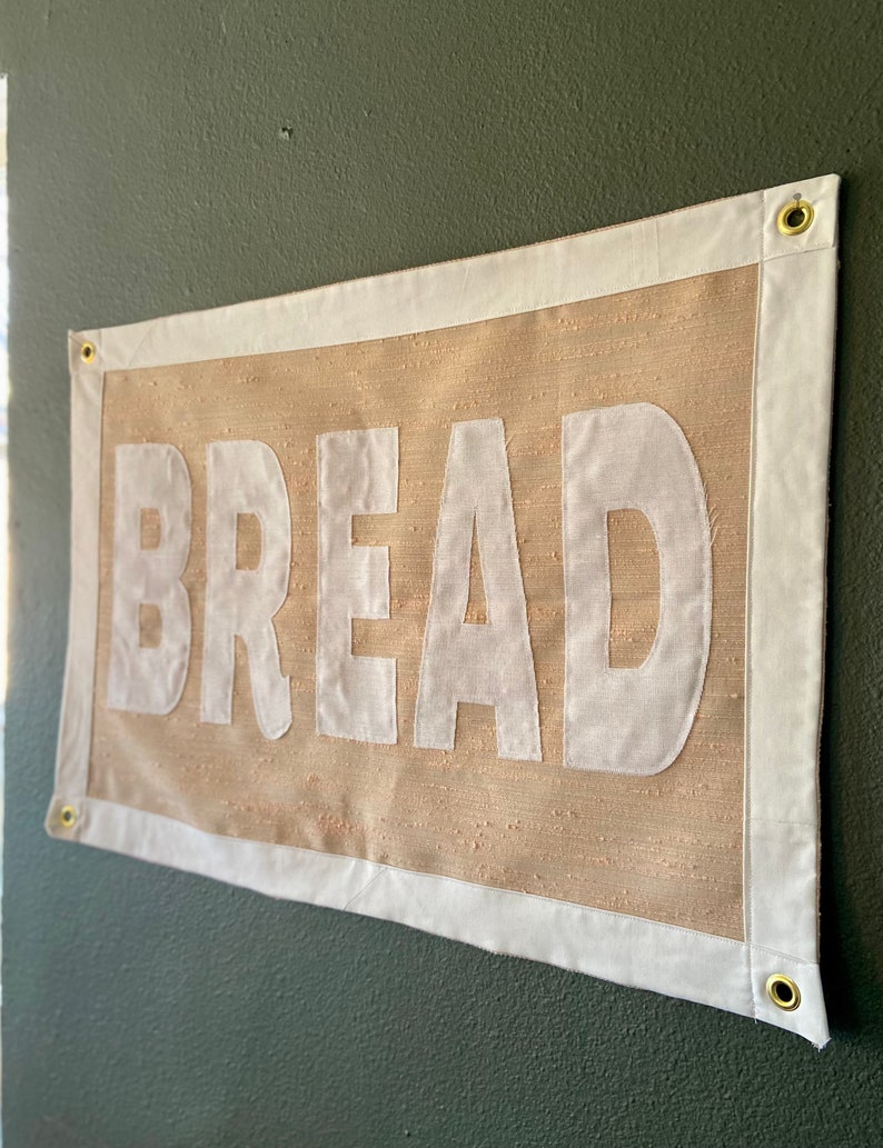 May include: A beige canvas banner with white trim and the word "BREAD" printed in large white letters.