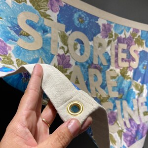 May include: A white canvas banner with a floral print in shades of blue, purple, and green. The banner has the words "Stories Are Fine" printed in white on it. The banner has a brass grommet in the corner.