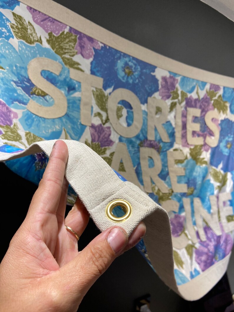 May include: A white canvas banner with a floral print in shades of blue, purple, and green. The banner has the words "Stories Are Fine" printed in white on it. The banner has a brass grommet in the corner.