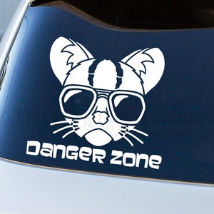 May include: White vinyl decal of a cat's face wearing sunglasses, with the text "DANGER ZONE" below. The cat has striped markings on its head and ears. The decal is applied to a car window.
