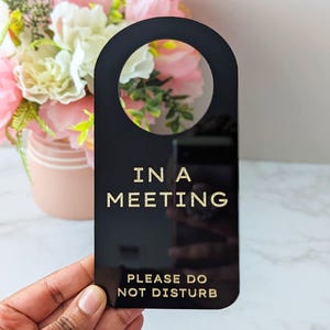 May include: Black door hanger with gold lettering reading "IN A MEETING PLEASE DO NOT DISTURB". The hanger has a rounded top with a circular cutout. A bouquet of pink and white flowers is in the background.