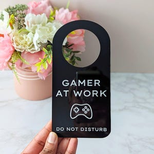 May include: Black door hanger with a circular top section. White text reads "GAMER AT WORK" and "DO NOT DISTURB", with a white controller graphic. A vase of pink and white flowers is in the background.