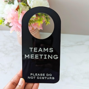 May include: Black door hanger with a circular cutout at the top. The text "TEAMS MEETING PLEASE DO NOT DISTURB" is etched in white. The background includes a vase of pink and white flowers.
