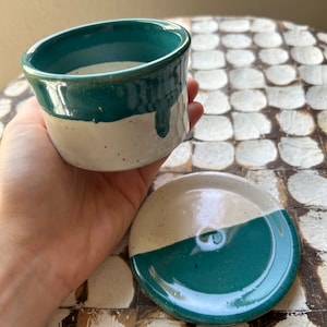 Puede incluir: Un juego de dos platos de cerámica, uno un tazón pequeño y el otro un platillo. Ambos son blancos con una banda verde azulada alrededor de la parte superior.