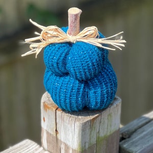 Op de afbeelding: Een tealblauwe gebreide pompoen met een strooien strik en een houten steel. De pompoen staat op een houten paal.