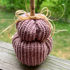 Op de afbeelding: Een gebreide pompoenversiering in mauve kleur met een taksteel en een strooien strik.