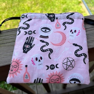 May include: Pink drawstring bag with a black and white print of various mystical symbols, including moons, stars, snakes, hands, skulls, and eyes. The bag is perfect for storing dice, tarot cards, or other small items.