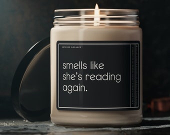 May include: A beige candle with a black label that reads "smells like she's reading again." The candle is lit and the flame is visible.