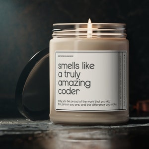 Puede incluir: Una vela de soja beige encendida en un tarro de cristal con tapa negra. La etiqueta dice "smells like a truly amazing coder". La vela está hecha a mano en California. Hecha con un poco de esfuerzo y mucho amor.