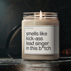 May include: A lit soy candle in a clear glass jar with a black lid. The label reads "smells like kick-ass lead singer in this b*tch". The candle is hand-poured in California.