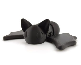 Könnte beinhalten: Eine schwarze Plastik-Fledermaus-Figur mit großen Ohren und Flügeln. Die Fledermaus liegt auf der Seite mit ausgebreiteten Flügeln.