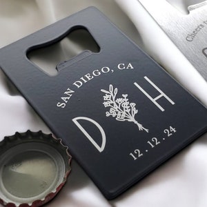 May include: Two rectangular bottle openers with a bottle cap. One is dark gray with white text and a floral design, the other is silver with text and champagne glasses. The dark gray opener says "SAN DIEGO, CA" and "D H 12.12.24". The silver opener says "Cheers to many years! GAR 04-24-2024".
