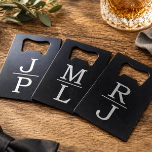 May include: Three rectangular black metal bottle openers, each featuring a cutout at the top and a personalized monogram design. The monograms consist of two white capital letters separated by a horizontal line, specifically J and P, M and L, and R and J.