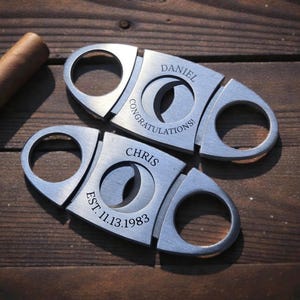 May include: Two silver-toned metal cigar cutters with finger loops. One cutter is engraved with the name DANIEL and the word CONGRATULATIONS. The other cutter is engraved with the name CHRIS and the text EST. 11.13.1983. A brown cigar is visible.