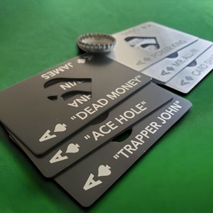 May include: A stack of rectangular metal bottle openers shaped like playing cards. The black cards feature white text including "DEAD MONEY," "ACE HOLE," and "TRAPPER JOHN," while the silver cards display "POKER KING," "MR. ALL-IN," and "CARD SHARK."
