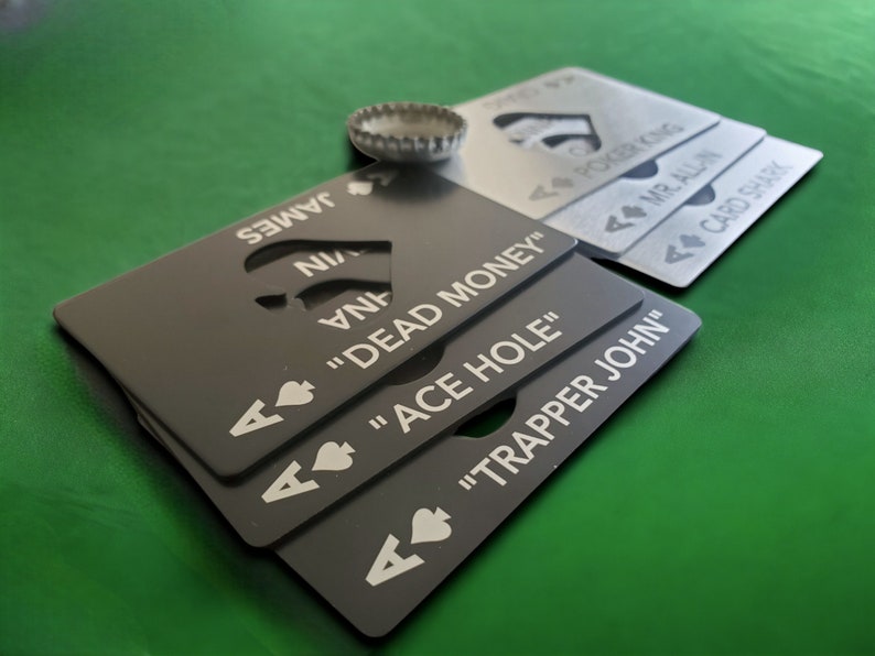 May include: A stack of rectangular metal bottle openers shaped like playing cards. The black cards feature white text including "DEAD MONEY," "ACE HOLE," and "TRAPPER JOHN," while the silver cards display "POKER KING," "MR. ALL-IN," and "CARD SHARK."
