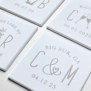 May include: Four white square coasters with gray text. Each coaster has a different design, including initials, dates, and locations like Raleigh, NC, and Big Sur, CA. The coasters are likely for a wedding or special event.