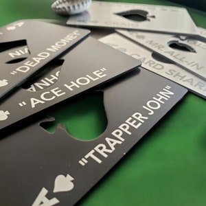 May include: A collection of rectangular metal bottle openers shaped like playing cards, featuring spade symbols and custom text including "DEAD MONEY," "ACE HOLE," and "TRAPPER JOHN." The openers are finished in matte black and brushed silver.