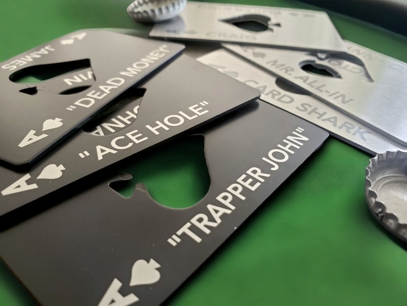 May include: A collection of rectangular metal bottle openers shaped like playing cards, featuring spade symbols and custom text including "DEAD MONEY," "ACE HOLE," and "TRAPPER JOHN." The openers are finished in matte black and brushed silver.