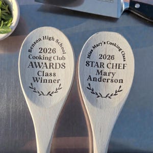 May include: Two wooden spoons with engraved text. One reads "Benson High School 2026 Cooking Club AWARDS Class Winner." The other says "Miss Mary's Cooking Class 2026 STAR CHEF Mary Anderson." A knife and a bowl of herbs are in the background.