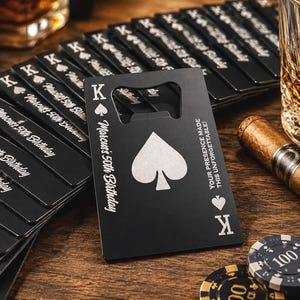 May include: A stack of matte black metal bottle openers shaped like playing cards. Each card features a white spade symbol, the letter K, and the text Marcus's 50th Birthday and Your Presence Made This Unforgettable engraved on the surface.