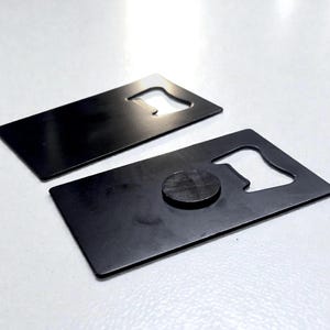 May include: Two rectangular, matte black metal bottle openers shaped like credit cards. Each opener features a cutout notch on one side for removing bottle caps. One of the openers has a small, circular black magnet attached to its surface.