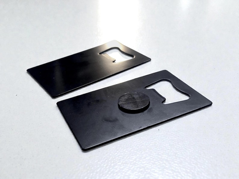 May include: Two rectangular, matte black metal bottle openers shaped like credit cards. Each opener features a cutout notch on one side for removing bottle caps. One of the openers has a small, circular black magnet attached to its surface.