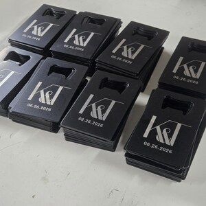 May include: A collection of rectangular black metal bottle openers arranged in stacks. Each opener features a laser-engraved design with the letters K&T and the date 06.26.2026 printed in a white font. The openers have a cutout at the top.