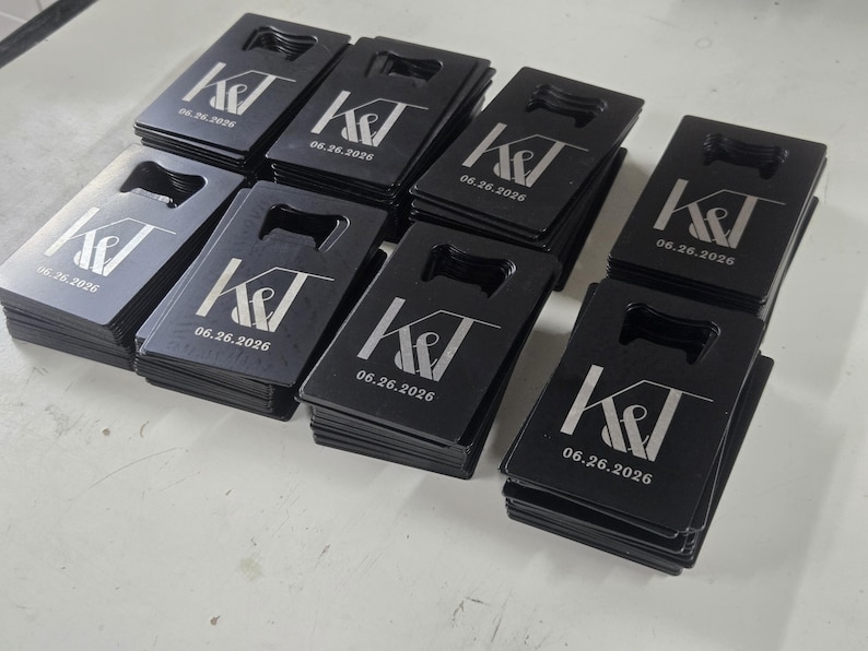 May include: A collection of rectangular black metal bottle openers arranged in stacks. Each opener features a laser-engraved design with the letters K&T and the date 06.26.2026 printed in a white font. The openers have a cutout at the top.