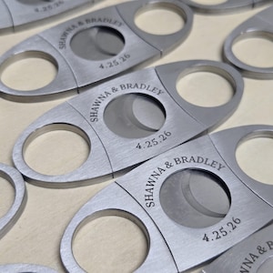 May include: A collection of silver-toned metal cigar cutters featuring a brushed finish. Each cutter has two circular finger holes and a central circular blade opening. The text SHAWNA & BRADLEY and the date 4.25.26 are engraved on the surface.