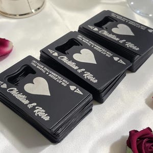 May include: Three stacks of matte black credit card-sized bottle openers shaped like playing cards. Each features an ace of hearts design, the names Christian & Kiera, and the date 06.27.2026 with the text FOR CELEBRATING WITH US engraved in white.