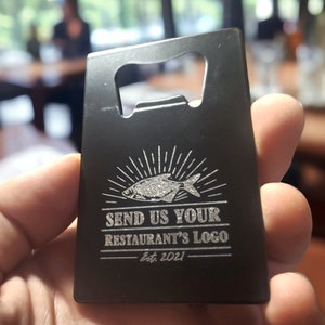 May include: A rectangular black metal bottle opener shaped like a credit card. The surface features an engraved graphic of a fish with sunburst lines above it, followed by the text "SEND US YOUR RESTAURANT'S LOGO" and "Est. 2021" below.