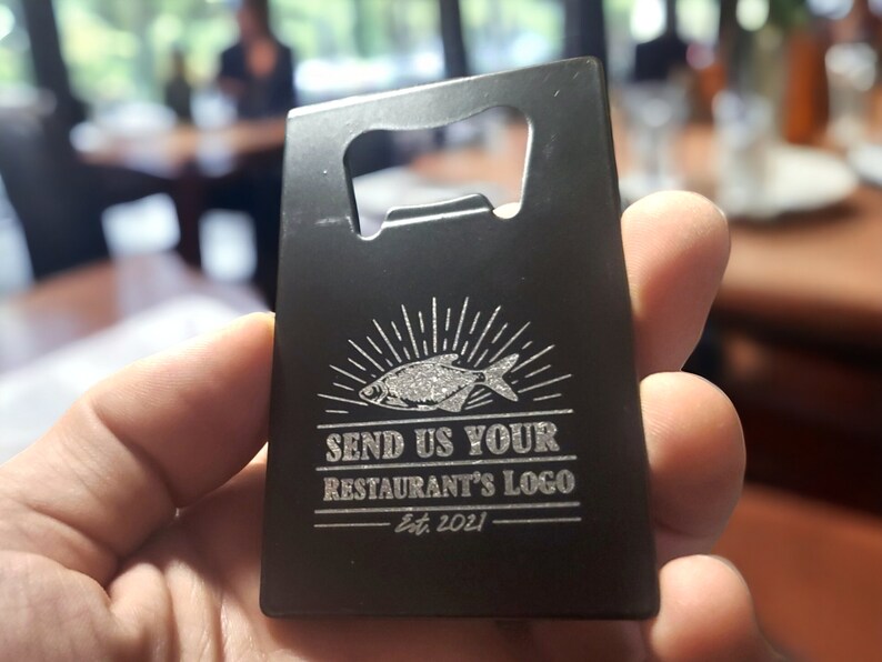 May include: A rectangular black metal bottle opener shaped like a credit card. The surface features an engraved graphic of a fish with sunburst lines above it, followed by the text "SEND US YOUR RESTAURANT'S LOGO" and "Est. 2021" below.