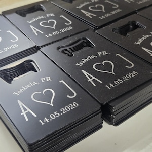 May include: Black bottle openers with a rectangular shape and a built-in opener. Each opener is engraved with the text "Isabela, PR", a heart symbol, the letters "A J", and the date "14.05.2026".