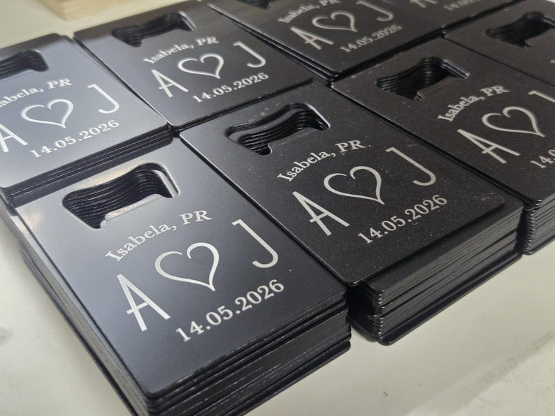 May include: Black bottle openers with a rectangular shape and a built-in opener. Each opener is engraved with the text "Isabela, PR", a heart symbol, the letters "A J", and the date "14.05.2026".