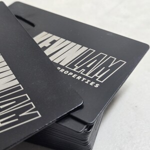 May include: A stack of rectangular black metal business cards with rounded corners. Each card features the silver-toned engraved text KEVIN LAM PROPERTIES in a bold, sans-serif font. The cards have a matte finish and a small cutout at the top.