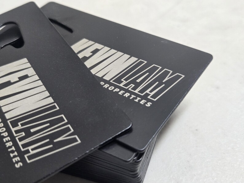 May include: A stack of rectangular black metal business cards with rounded corners. Each card features the silver-toned engraved text KEVIN LAM PROPERTIES in a bold, sans-serif font. The cards have a matte finish and a small cutout at the top.