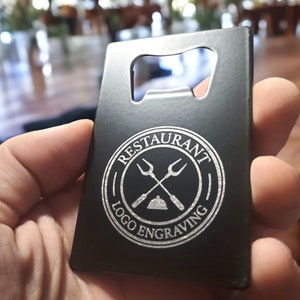 May include: A rectangular black metal bottle opener shaped like a credit card. The surface features a circular white engraved logo with crossed forks and a serving dome, surrounded by the text RESTAURANT LOGO ENGRAVING in a circular border.