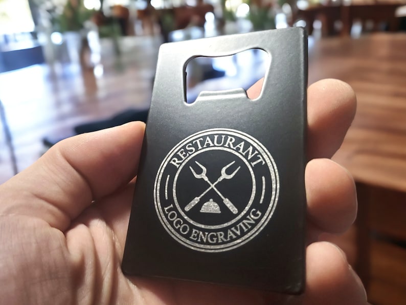 May include: A rectangular black metal bottle opener shaped like a credit card. The surface features a circular white engraved logo with crossed forks and a serving dome, surrounded by the text RESTAURANT LOGO ENGRAVING in a circular border.