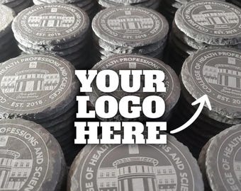 May include: A collection of circular dark gray slate coasters featuring an engraved building illustration, the text College of Health Professions and Sciences, Est. 2018, and Five-year Anniversary. The image includes the text YOUR LOGO HERE.